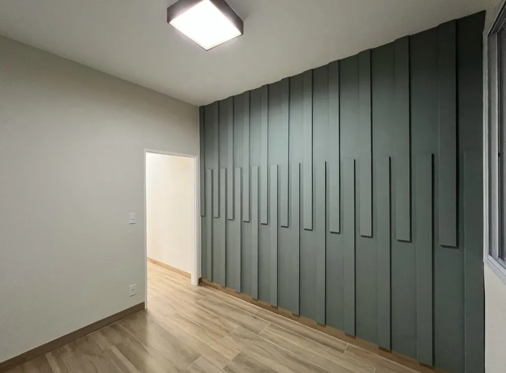 Quarto de anexo residencial com painel ripado geométrico na cor verde-oliva cobrindo a parede de destaque, piso amadeirado e teto com luminária plafon quadrada de sobrepor.