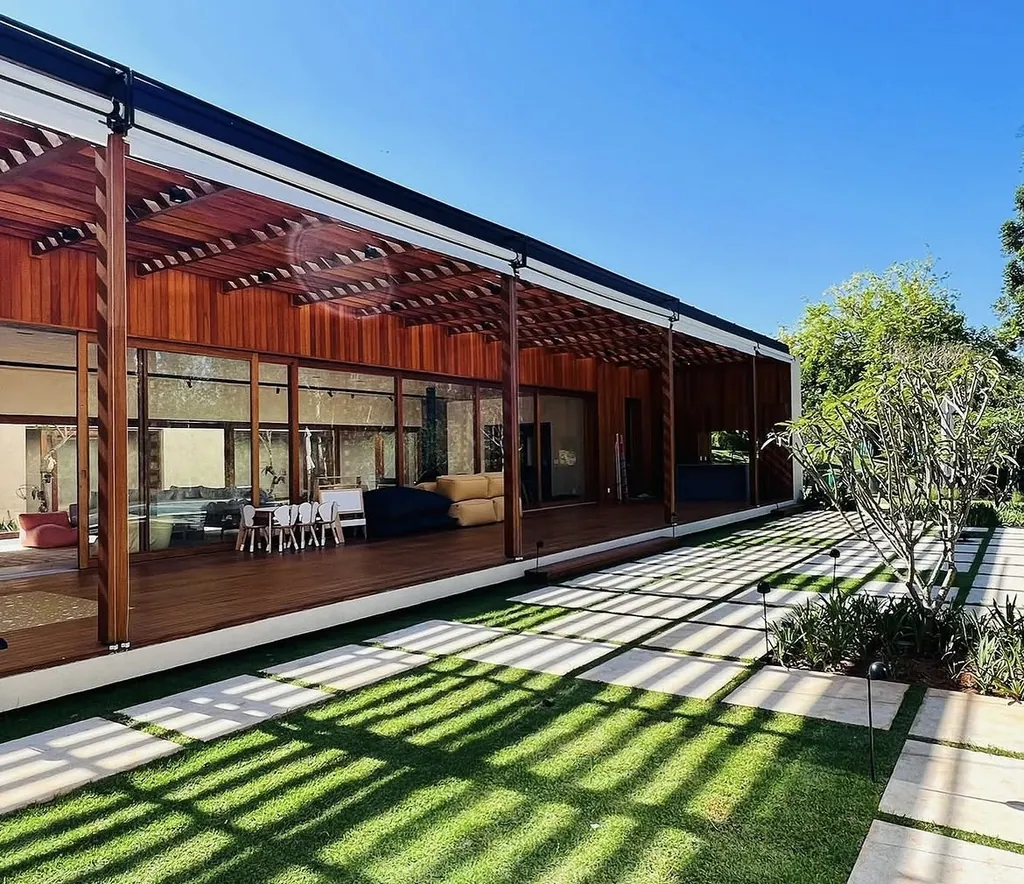 Varanda gourmet de residência de alto padrão com pérgola e revestimento de madeira no teto, grandes portas de vidro de correr e piso em deck. À frente, gramado com placas de pedra e sombras projetadas sob céu azul.