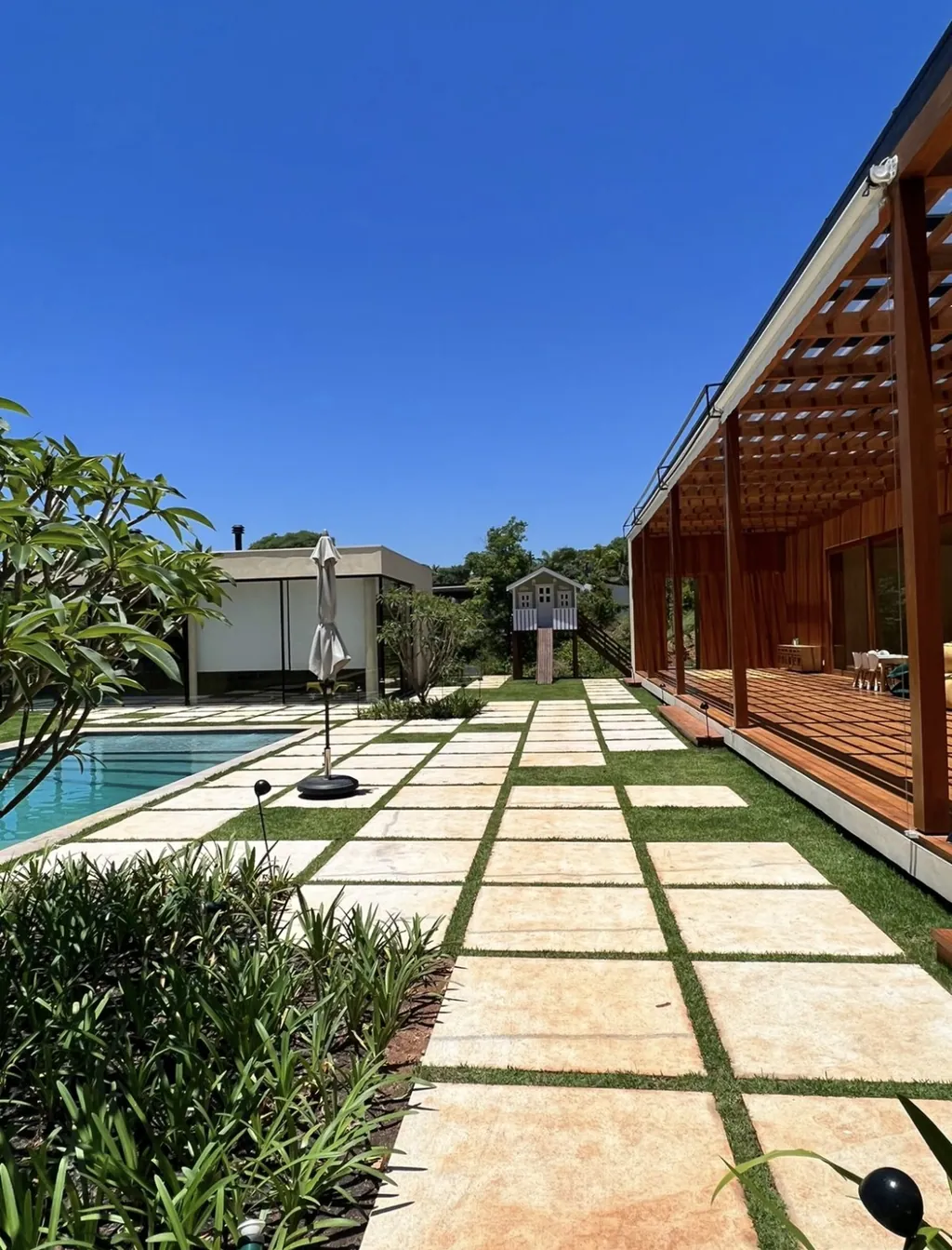 Vista longitudinal do jardim de uma casa moderna, mostrando caminho em placas de pedra sobre o gramado, piscina ao fundo à esquerda, grande varanda com cobertura de madeira à direita e casinha infantil ao fundo.